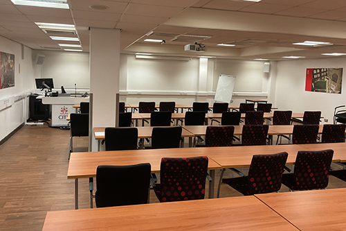 Lecture room with multiple rows of tables and chairs facing a whiteboard with overhead projector, and presentation PC podium.